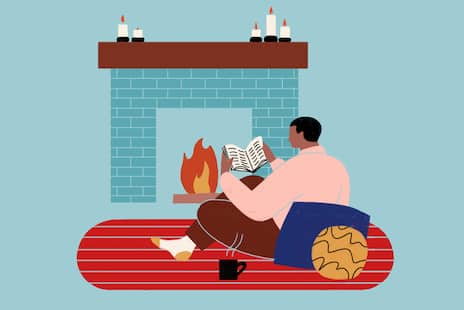An illustration of a man reading a book by the fire, sitting on a red rug, leaning against some pillows. There are candles on the mantle and a steaming mug next to him.